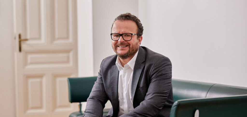 Rechtsanwalt Dr. Kopecky sitzt im Anzug lächelnd auf einem Sofa.