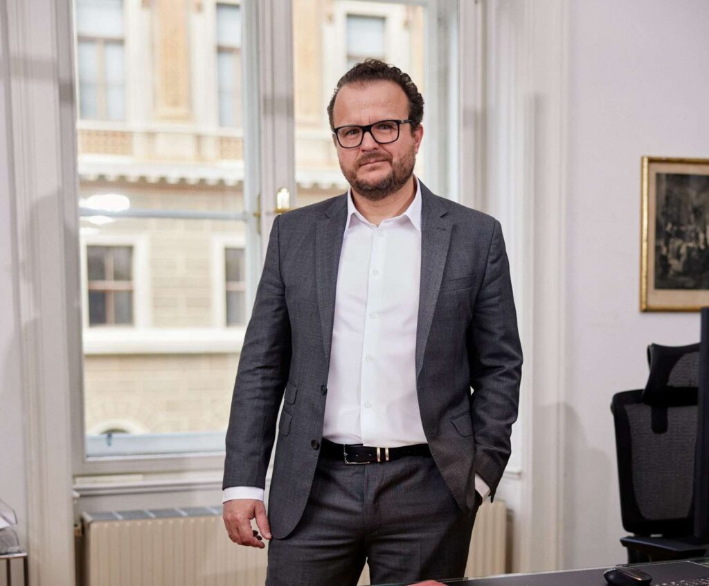 Dr. Kopecky, Rechtsanwalt, steht im Anzug in einem Büroraum vor einem Fenster mit Blick auf ein historisches Gebäude.